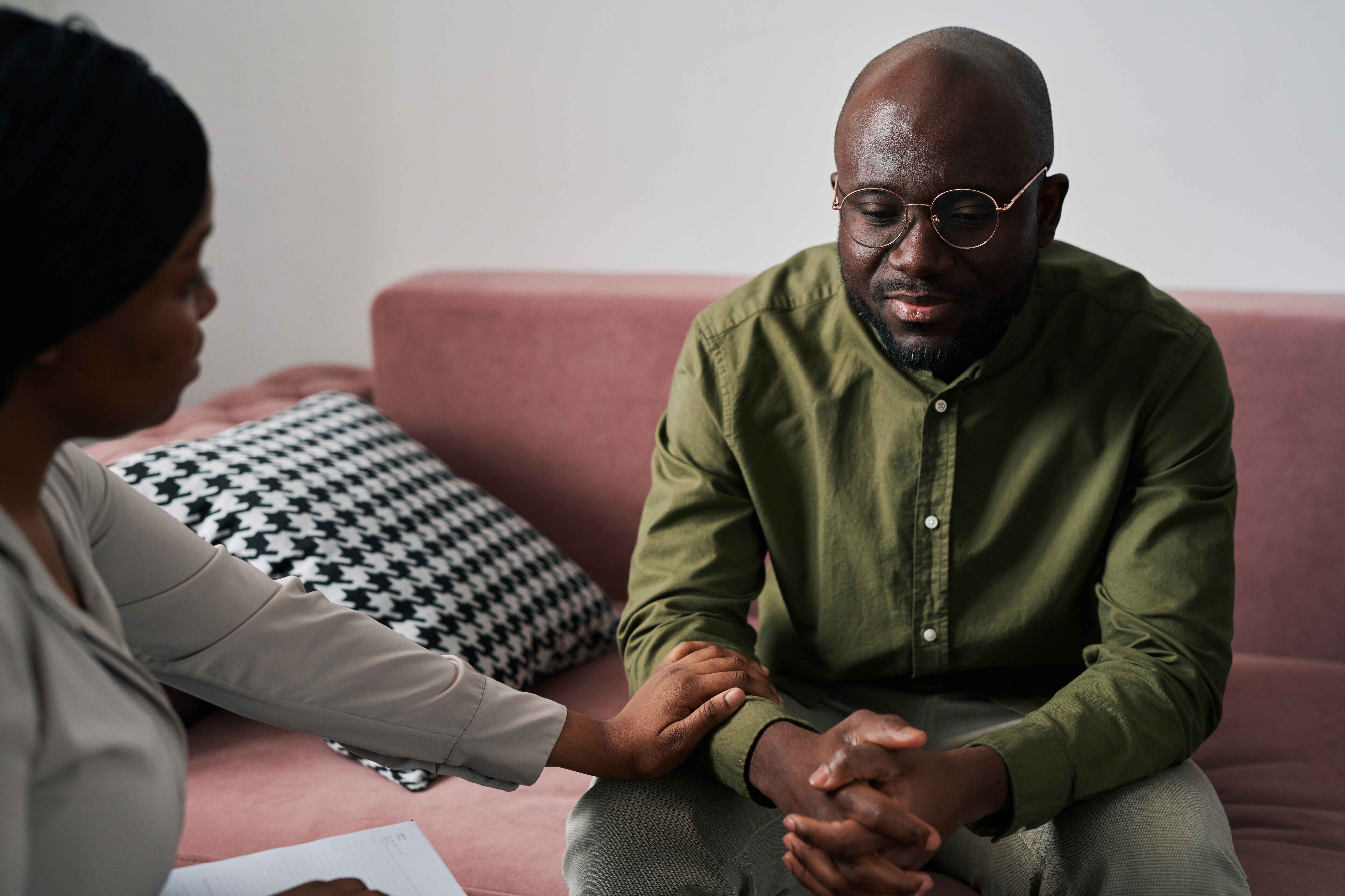 Man sitting on a couch, appearing contemplative and engaged in therapy session, with a therapist's hand gently resting on his arm, emphasizing emotional support in trauma-focused psychotherapy.